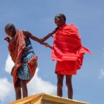 Two Maasai warriors clown around. They are dressed in traditional clothing including bright red shuka rugs.